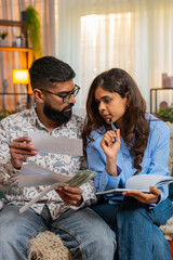 Indian couple man and woman sitting on home sofa, calculating bills and expenses with money and notebook, showing concern. Family managing home budget as girl writes and man reacts with frustration.