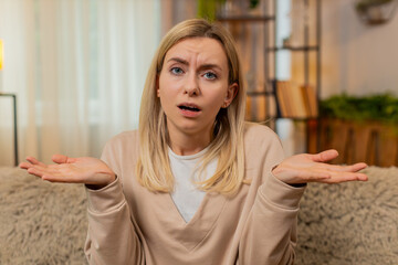 Young adult woman spreads hands shakes head while sitting on home sofa, reacting to negative online...
