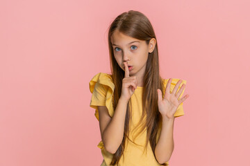 Young child girl smiles gently while placing a finger on lips, making a shh gesture suggesting to be quiet or keep a secret. School kid isolated on pink background playful and expressive communication