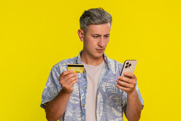 Middle-aged man holding smartphone and credit card while attentively entering payment information to make online purchase shopping. Young guy concentrated on transaction isolated on yellow background.