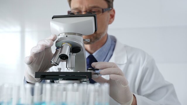 Scientific researcher wearing lab coat, glasses and white protective gloves carefully calibrating laboratory microscope, performing precision medical investigation. Medicine, healthcare and science