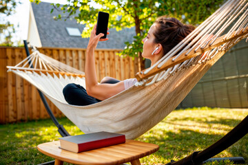 Woman with earbuds relaxing in a hammock using a smartphone outdoors.