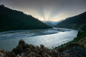 Teesta river flowing through Himalayan mountains, sunlight coming through clouds in the background, setting sun behind mountain peaks. Sikkim, India.