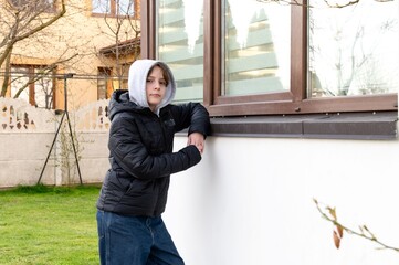 Young boy in black jacket leans against a wall in a backyard during early afternoon