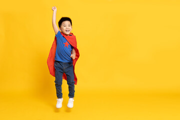 Asian little boy superhero smiling, hand up and jumping in red cape isolated yellow background, Full body composition
