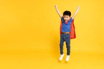 Asian little boy superhero smiling, hand up and jumping in red cape isolated yellow background, Full body composition
