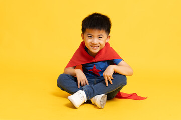 Asian little boy superhero smiling and sitting on floor in red cape isolated yellow background, Full body composition