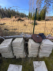 Square white tiles are stacked in three piles on grass near a wire fence, with a rusted metal object and black bag on one pile. Sparse trees and dry grass extend beyond.