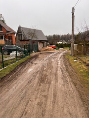 A muddy dirt road passes through a residential area with a wooden house, a brick house under construction, a red car, and sparse trees under overcast skies.