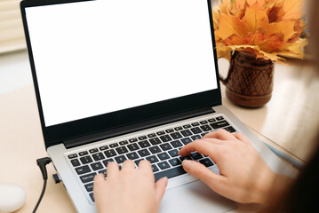 Hands typing on laptop keyboard with blank white screen and autumn leaves decoration on desk.