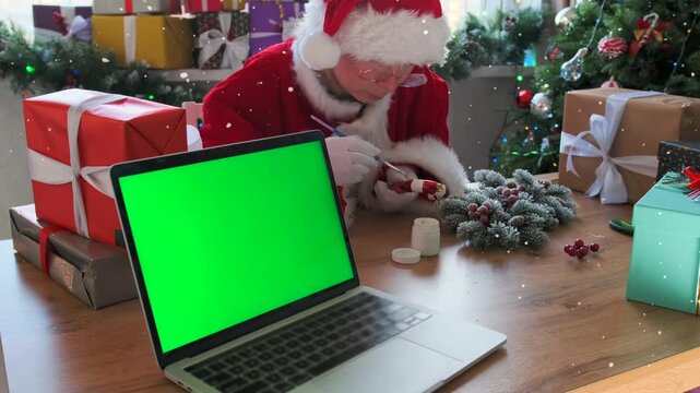 Woman in festive costume paints a red and white candy cane decoration by hand while sitting at a wooden table with wrapped gifts and holiday wreath. A laptop with green screen is placed nearby.
