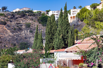 Terraced construction of houses with red tile roofs on the mountain slopes of Javea, with landscaping of cypress trees and flowering shrubs, Spain