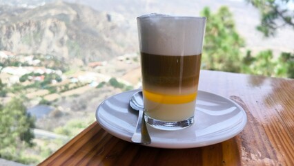 Traditional Canary Islands coffee Barraquito served on a wooden table at a mountain café in Gran Canaria, Spain, with scenic view of green hills and valleys in soft daylight atmosphere.