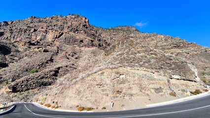 Fototapeta premium Curved mountain road on Gran Canaria, Spain, surrounded by rugged volcanic cliffs and dry rocky slopes under clear blue sky. Typical dramatic highland landscape of the Canary Islands.