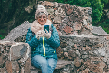 Smiling senior woman wearing winter clothes sitting outdoors using a smartphone. Concept of active elderly lifestyle, technology, communication, and enjoying nature.