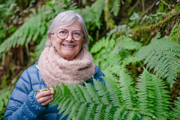 An active and smiling senior woman enjoys the freedom of the outdoors on a winter day, walking in a mountain forest touching a fern bush