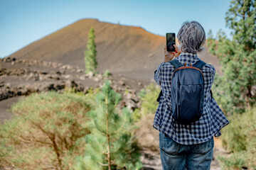 Rear view of elderly woman standing outdoors taking a picture to volcanic landscape in Tenerife island. Mountain, forest and blue sky on background