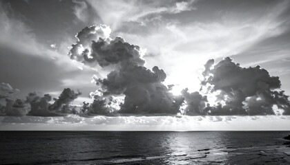 A grayscale coastal scene where clouds dominate the upper frame, lit by the sun peeking through. The ocean is in the mid, and a blurred beach below