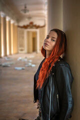 Obraz premium A beautiful girl near the doors of an abandoned palace. A girl with red hair. The beautiful interior of an abandoned palace. A leather jacket.