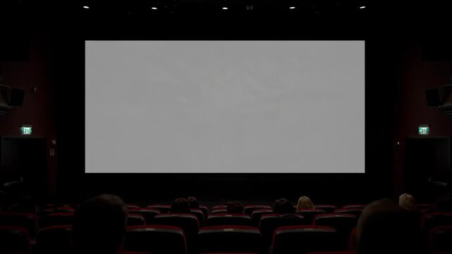 Empty Movie Screen in a Dark Cinema - A wide shot captures an empty movie screen in a dimly lit theater. Rows of red seats are filled with an audience waiting for the film to start.