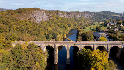 Drone shot of the impressive stone viaduct in Clécy, Normandy, spanning the Orne River. Framed by autumnal trees, the bridge creates a picturesque landscape amidst rolling hills and forests.