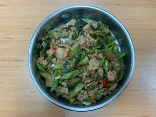 Stir-fried basil pork with chili on wooden table, green beans, and leafy greens in a stainless steel bowl—spicy, savory, and home-style Thai flavor