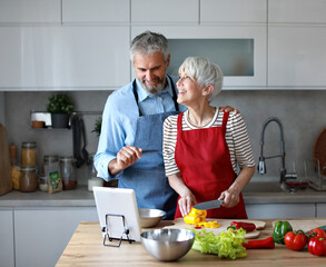 Portrait of happy senior mid aged mature couple prepering meal with fresh vegatebles and following internet instructions for a recipe on a tablet computer  or looking at video  or website app in kitch