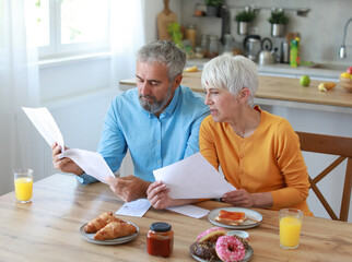 Senior couple checking bills and calculating expenses using a laptop during a breakfast in the morning at table at home, debt taxes concept, worried married elderly couple