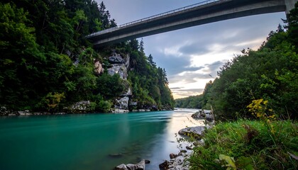 A long-exposure shot depicts a turquoise river flowing beneath a bridge. Lush green foliage and rocky cliffs frame the scene under a cloudy sky