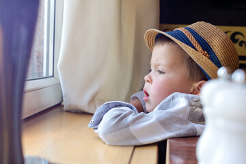 A little boy in a hat is leaning on the windowsill and looking out into the street. A child in a plaid shirt and hat looks out of an open window.