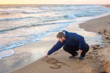 A man kneels on the beach, drawing with his finger in the sand next to gentle waves