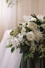 Floral wedding arrangement with white roses, greenery and soft light on beige curtain background. Elegant banquet hall decor. Concepts: celebration, romance, wedding, luxury. 