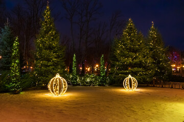 Large glowing balls lie near the Christmas trees decorated with garlands with lights. Festive decoration of the garden or park