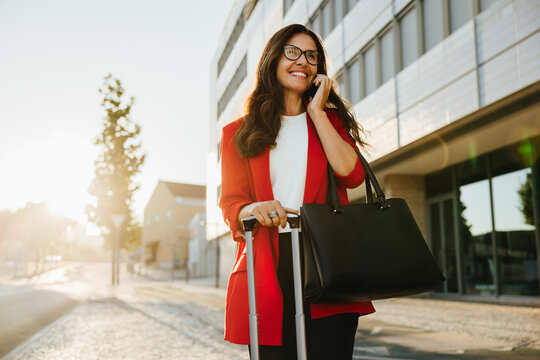 Fototapeta Smiling woman in red blazer carrying luggage and talking on phone outdoors