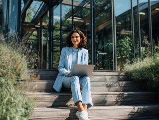 Confident businesswoman working on laptop outdoors with modern glass building background