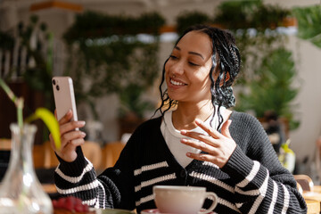Beautiful smiling African American woman having video call chatting on smartphone in cafe