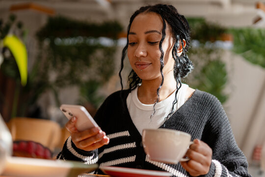 Smiling cheerful African American woman enjoying coffee holding mobile phone using mobile app - Powered by Adobe