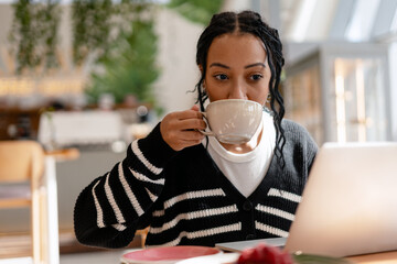Beautiful African American woman with braids and piercing drinking coffee using laptop in cafe