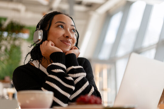 Fototapeta Young smiling African American woman student while listening to music in cafe using laptop