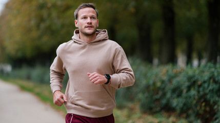 Young man in beige hoodie and maroon pants wearing a smartwatch running along a tree-lined path in city park