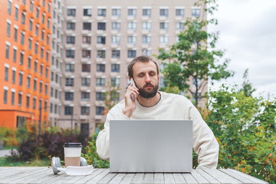Man working remotely outdoors with a laptop, talking on the phone, and having coffee, concept of remote work, digital nomad lifestyle, productivity, and communication