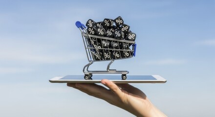 Hand holding digital tablet with miniature shopping cart filled with percentage symbol blocks against a clear blue sky