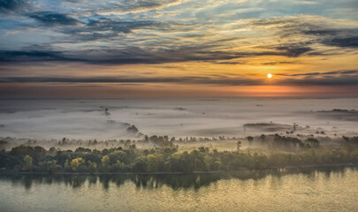 Autumn dawn over the river, where mist and clouds merge. The first rays of sunlight paint the water gold, capturing nature’s quiet awakening in a serene aerial view.