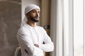 Thoughtful millennial middle eastern businessman stand by window at office