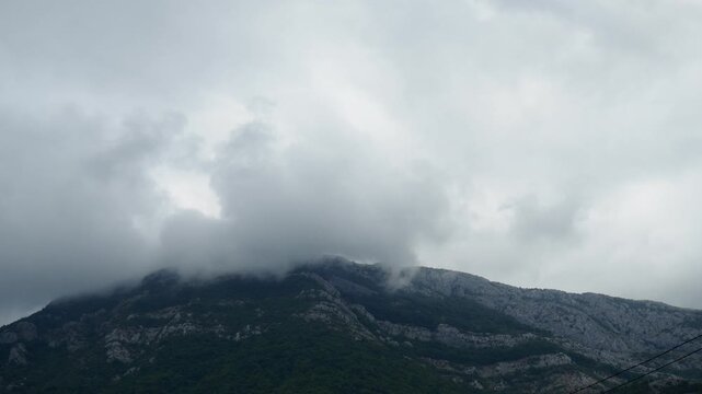 Overcast weather blankets the mountain range as thick clouds cling to the rocky peaks, creating a mysterious atmosphere. Lush greenery adds to the natural beauty. Montenegro, Canj, Bar