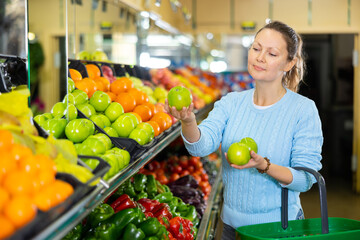 Obraz premium Delighted middle-aged woman customer buying fresh apples at the counter in big greengrocery