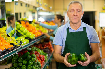 Positive aged salesman holding fresh big peppers at food stall in grocery store