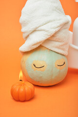 Halloween candle and pumpkin with eyes and a white terry towel on the head, a blue clay mask on the face, orange background