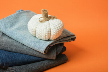 Decorative pumpkin and stack of jeans of different colors on orange background, autumn clothing sale concept