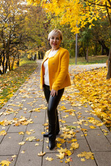 Fototapeta premium woman with short blonde hair stands in a park surrounded by autumn leaves. She wears a bright yellow jacket and black pants.